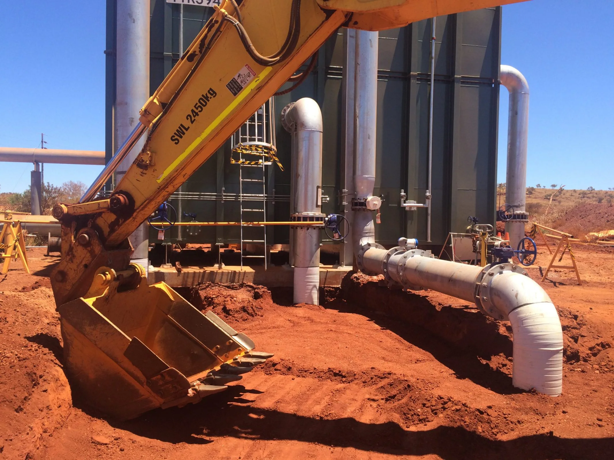 Yellow excavator digging dirt in front of large tank at civil construction site.